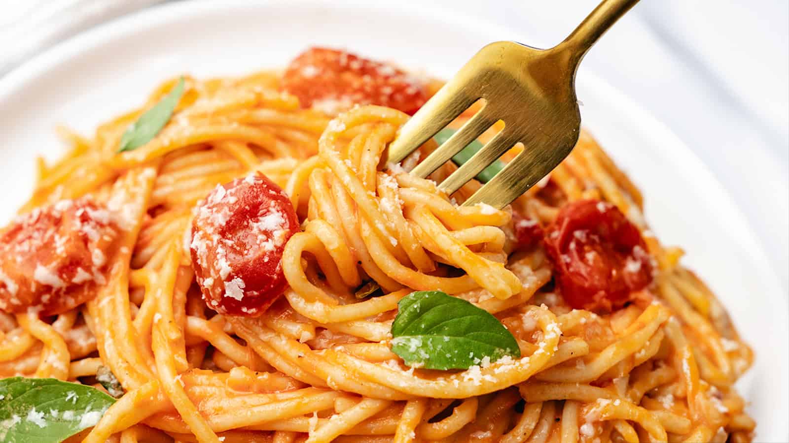 A fork twirling spaghetti with tomato sauce, cherry tomatoes, basil leaves, and grated cheese on a white plate.