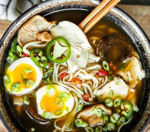 A bowl of ramen containing broth, noodles, sliced mushrooms, soft-boiled eggs, jalapeño slices, green onions, tender pieces of chicken, with chopsticks resting on the bowl's edge.