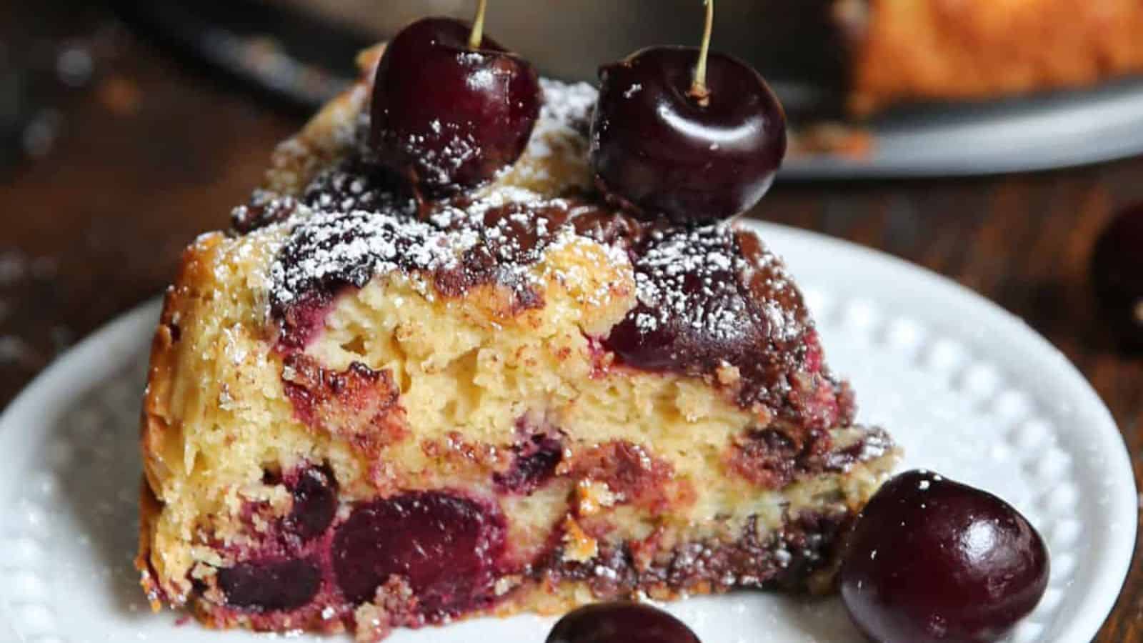 A slice of cherry cake topped with whole cherries and dusted with powdered sugar on a white plate.