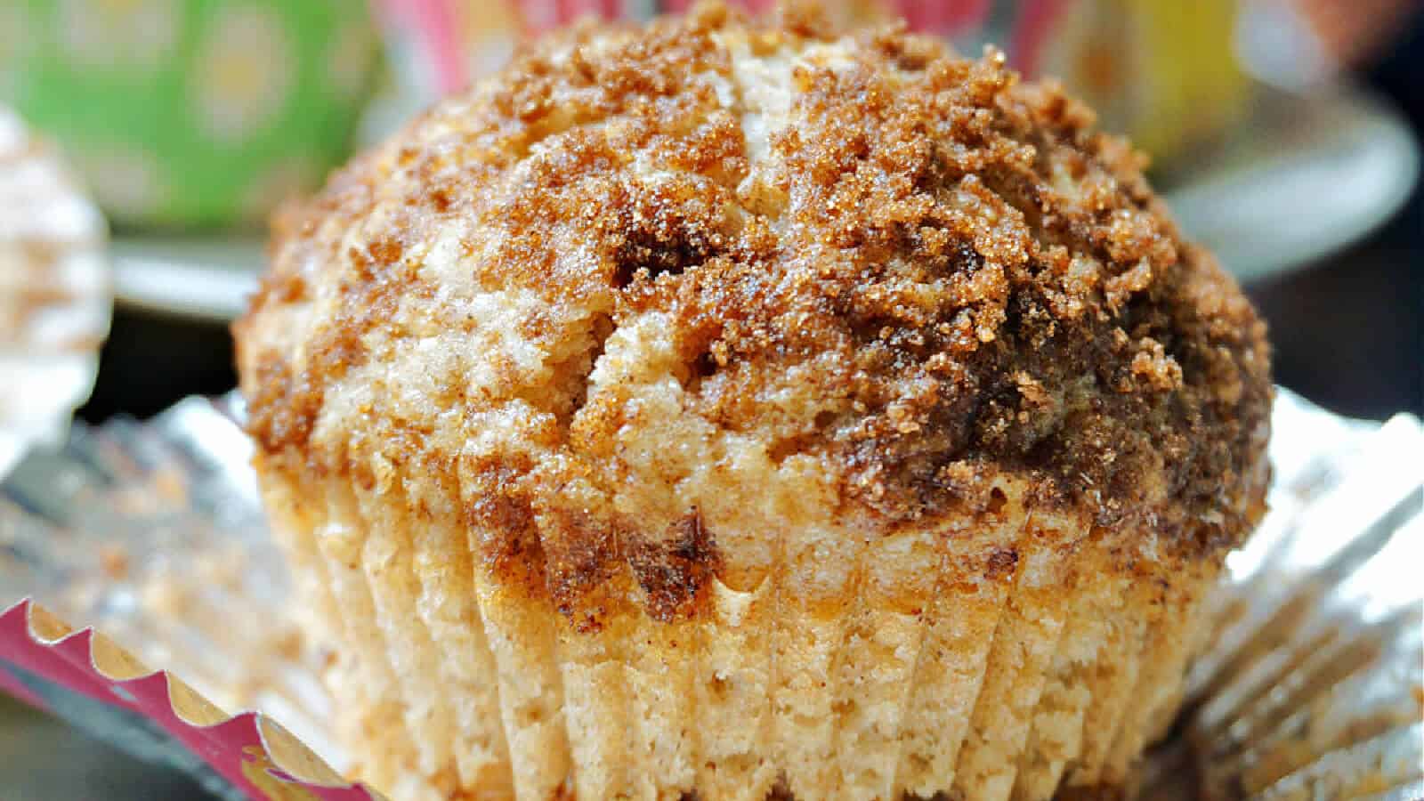 A close-up of a freshly baked muffin in an unwrapped paper liner. The muffin has a golden brown, crumbly texture on top, suggesting a streusel topping. The background is slightly blurred, featuring colorful, out-of-focus objects.