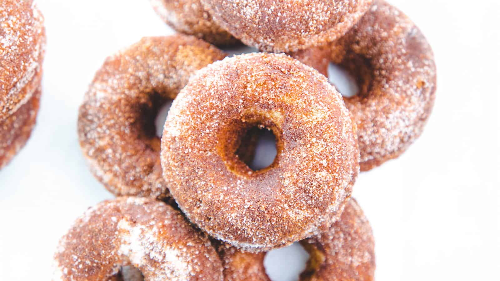 A close-up view of several stacked cinnamon sugar donuts. The donuts are golden-brown with a generous coating of cinnamon and sugar, creating a textured, sugary crust. The white background highlights the rich color and sugar coating of the donuts.