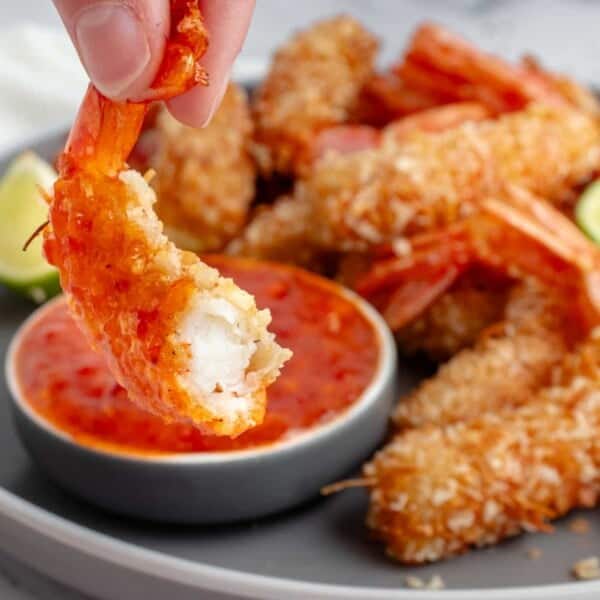 A hand holds a breaded shrimp with a bite taken out of it above a plate containing more fried shrimp, lime wedges, and a small bowl of orange dipping sauce.