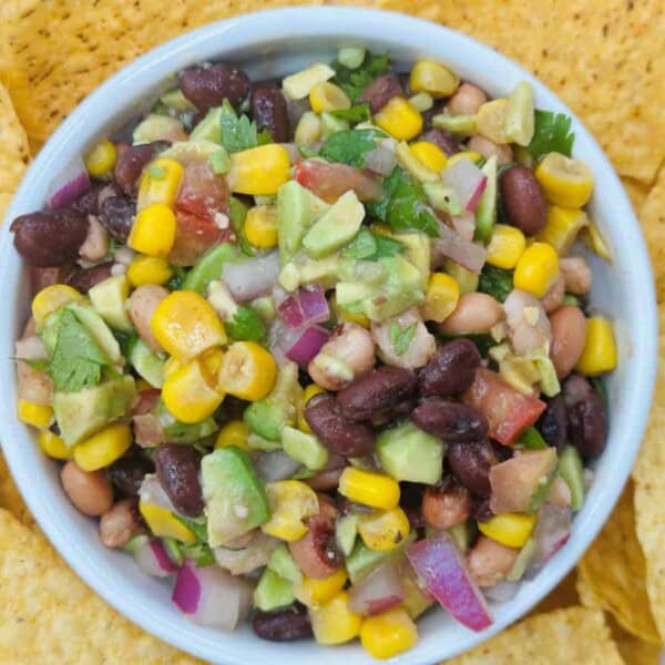 The image shows a bowl of salad with black beans, corn, avocado, red onion, and cilantro. Surrounding the bowl are crispy tortilla chips.