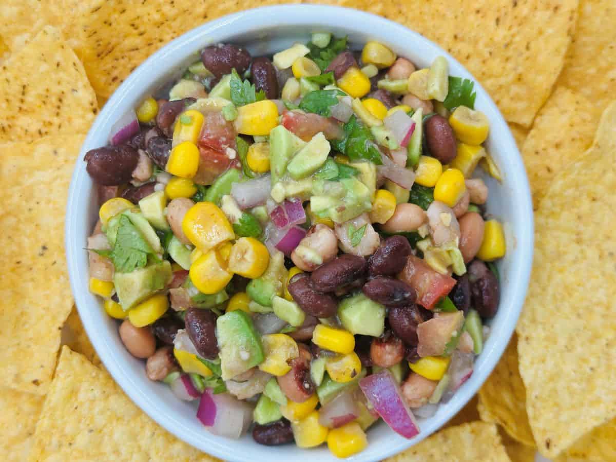 The image shows a bowl of salad with black beans, corn, avocado, red onion, and cilantro. Surrounding the bowl are crispy tortilla chips.