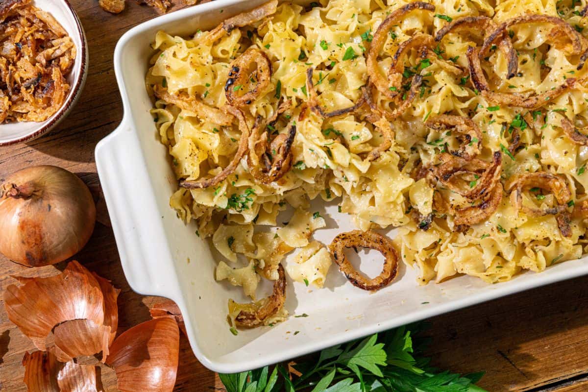 A casserole dish filled with noodle casserole, topped with crispy fried onions and parsley, sits on a wooden table next to a whole onion and fresh parsley.