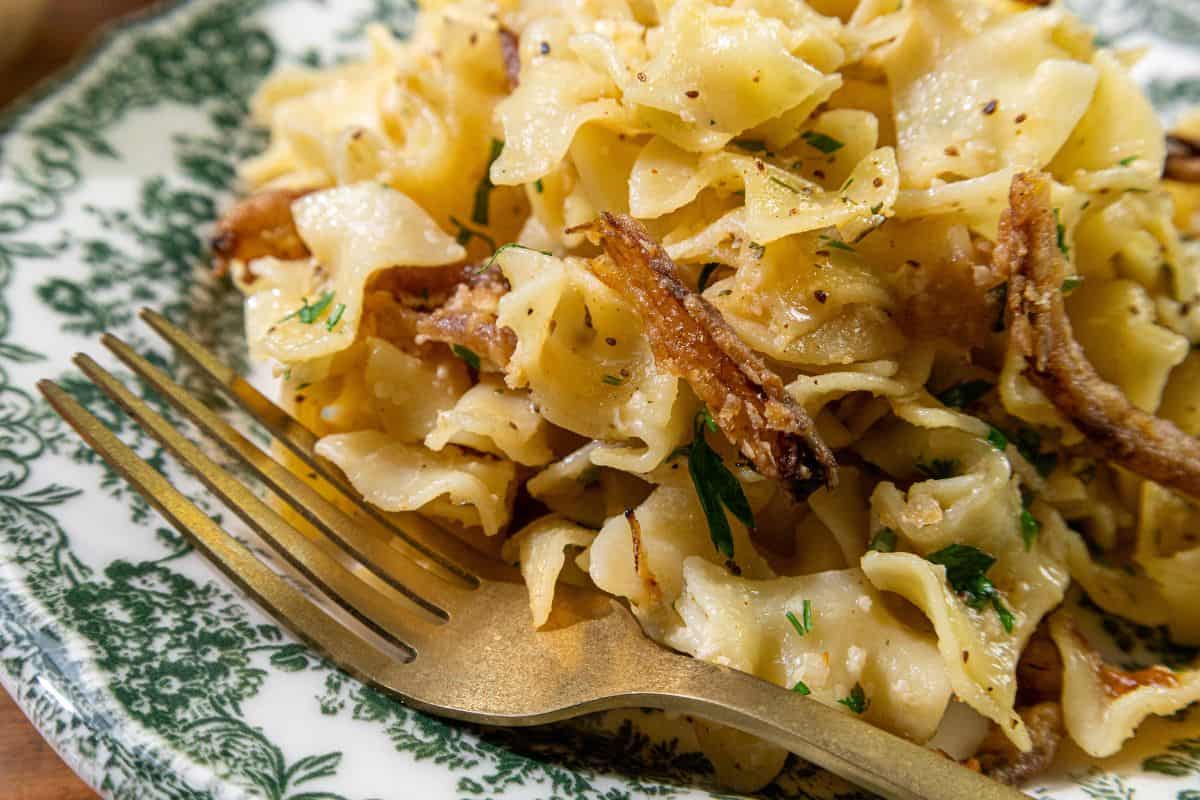 A plate of buttered egg noodles topped with caramelized onions and garnished with herbs, accompanied by a gold fork.