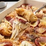 A fork lifting a piece of cheesy croissant breakfast casserole with sliced ham and apples in a baking dish.