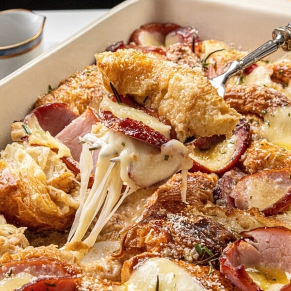 A fork lifting a piece of cheesy croissant breakfast casserole with sliced ham and apples in a baking dish.