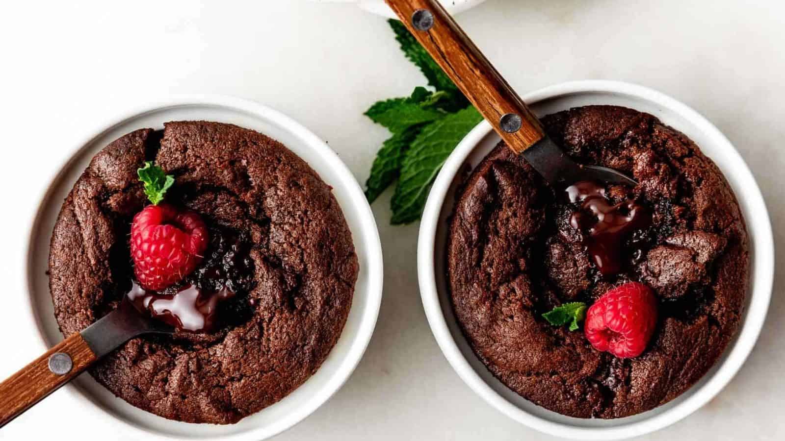 Two chocolate lava cakes in white ramekins, each topped with a raspberry and a mint leaf. Dessert spoons are inserted into the cakes, revealing melted chocolate centers.