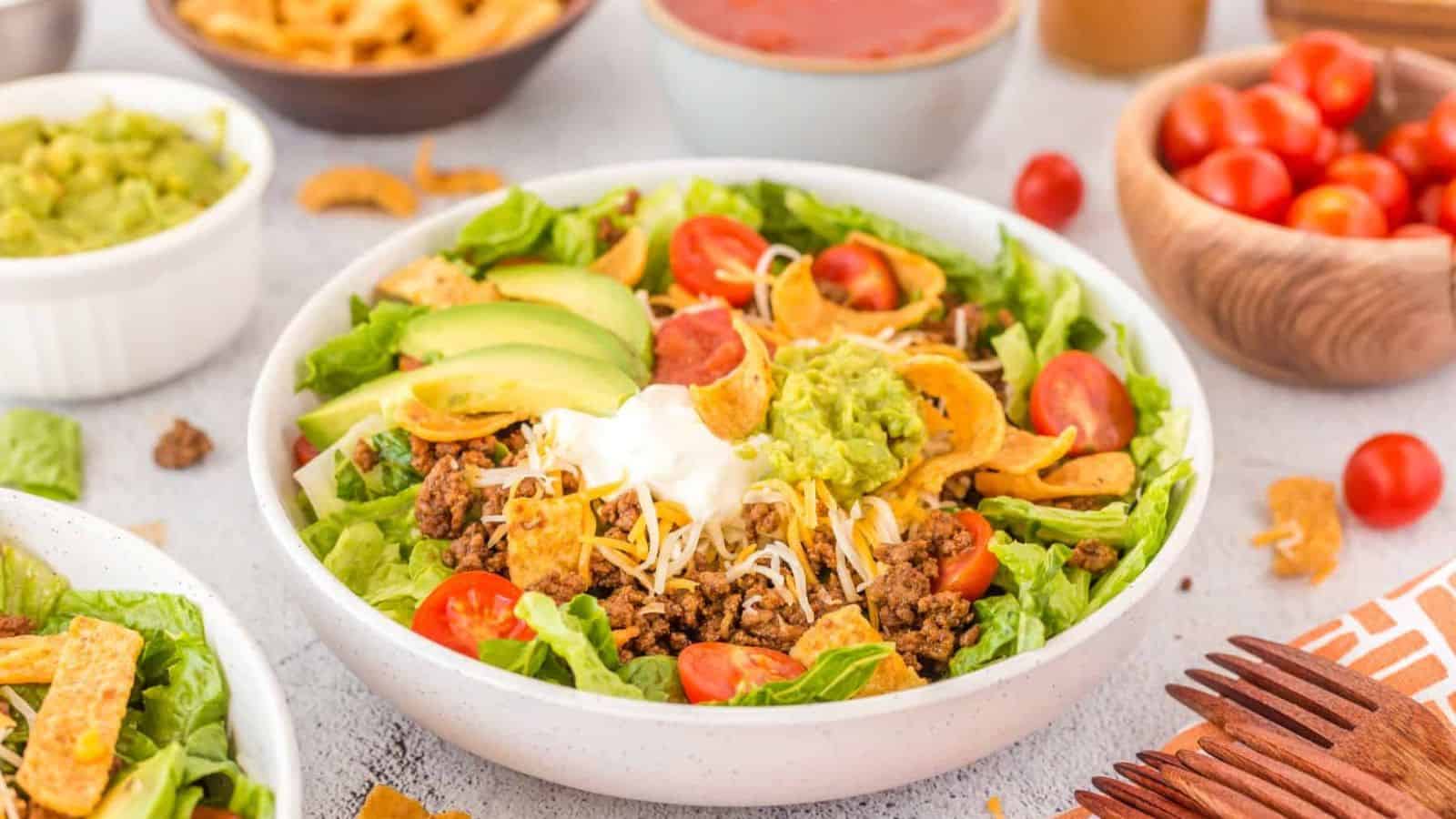 A taco salad in a white bowl with ground beef, lettuce, avocado slices, cherry tomatoes, shredded cheese, sour cream, and guacamole. It is surrounded by bowls of various side dishes.
