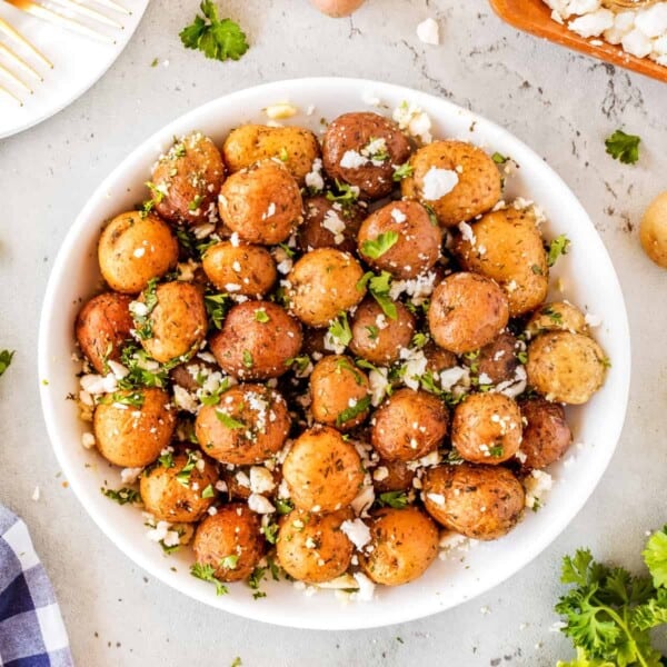 A white bowl filled with seasoned and roasted baby potatoes, garnished with chopped parsley and crumbled feta cheese. The bowl is placed on a light-colored surface, with scattered bits of parsley and feta around it.