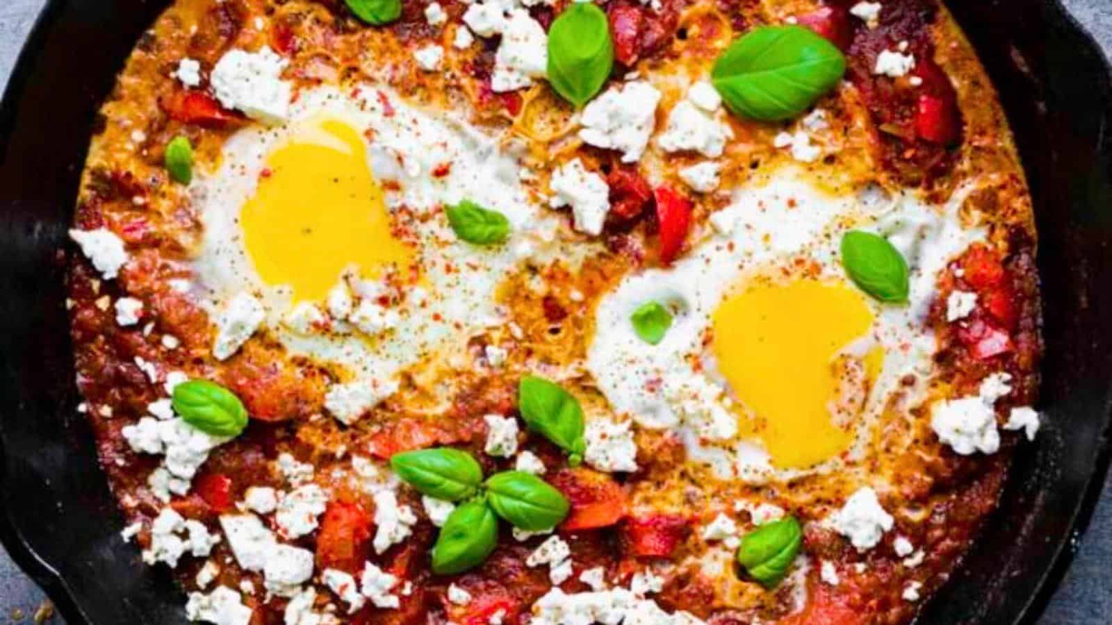 A black skillet containing shakshuka with two poached eggs, crumbled cheese, tomato sauce, and fresh basil leaves.