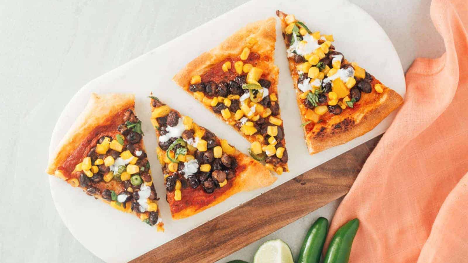 Four slices of vegetable pizza topped with corn, black beans, scallions, and a drizzle of white sauce, presented on a marble and wood tray with an orange cloth and some jalapeños nearby.