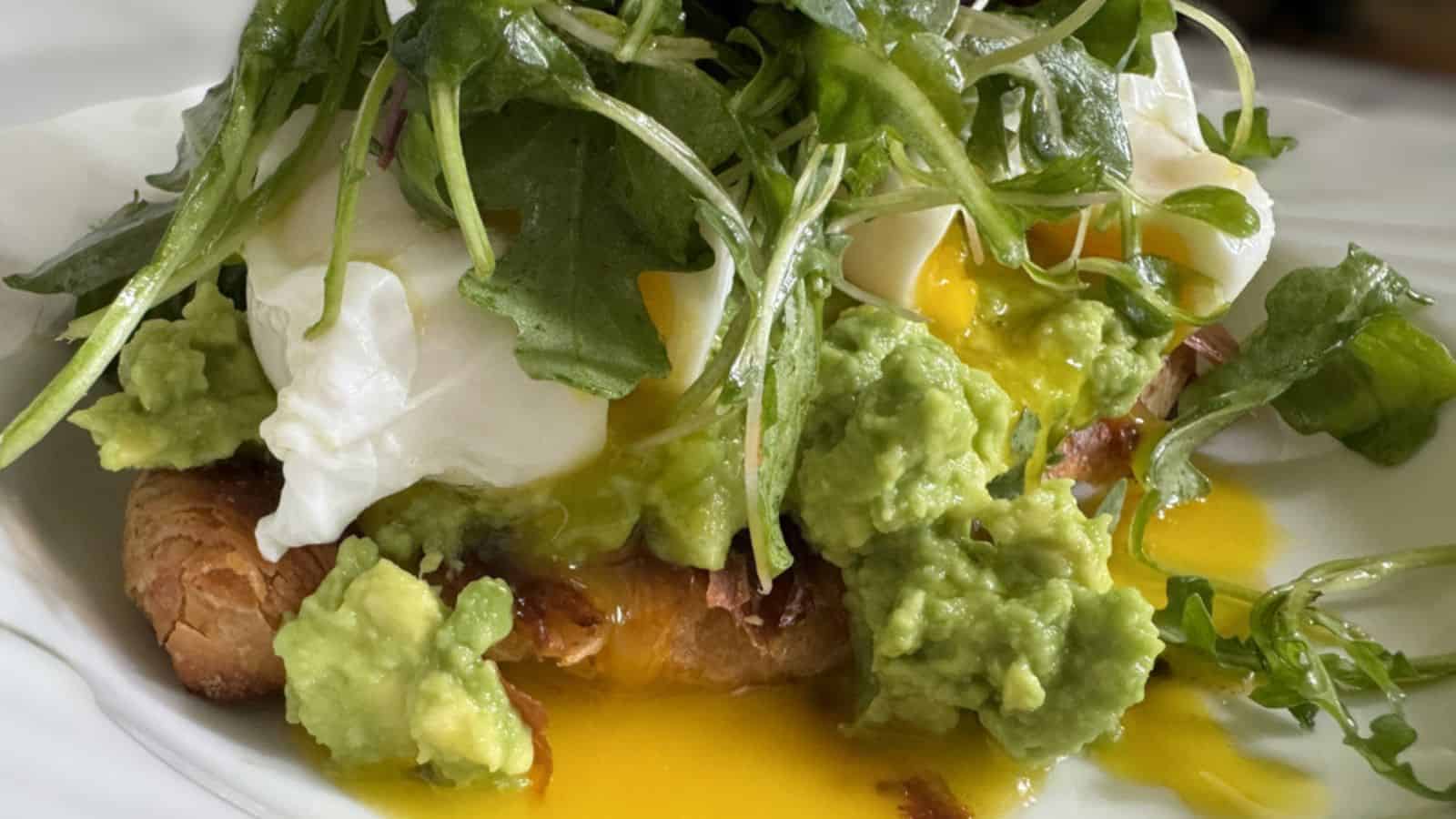 A close-up of a dish featuring poached eggs, mashed avocado, and greens on a slice of bread, with egg yolk running over the ingredients.