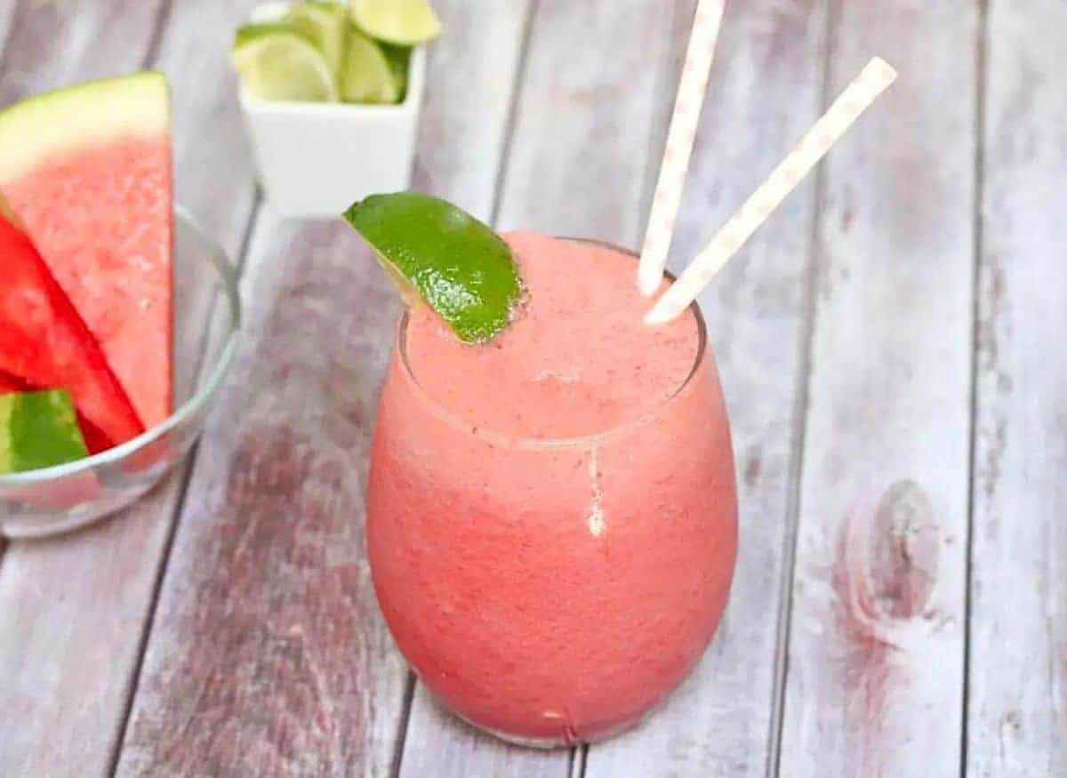 Pink watermelon slushie in a glass garnished with lime and more watermelon and lime behind it.