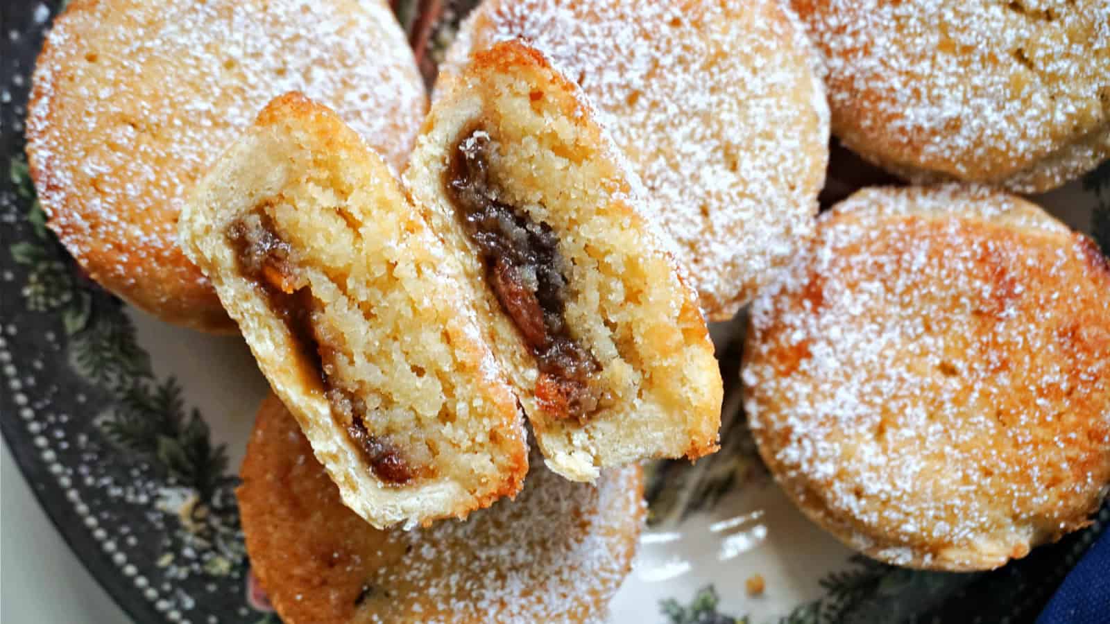 A close-up of several mince pies on a decorative plate. One pie is cut open to show the sweet filling inside. The pies are sprinkled with powdered sugar.