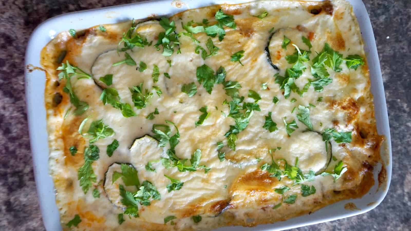 A casserole dish with visible layers of ground beef, melted cheese, fresh chopped herbs sprinkled on top, and slices of zucchini underneath.