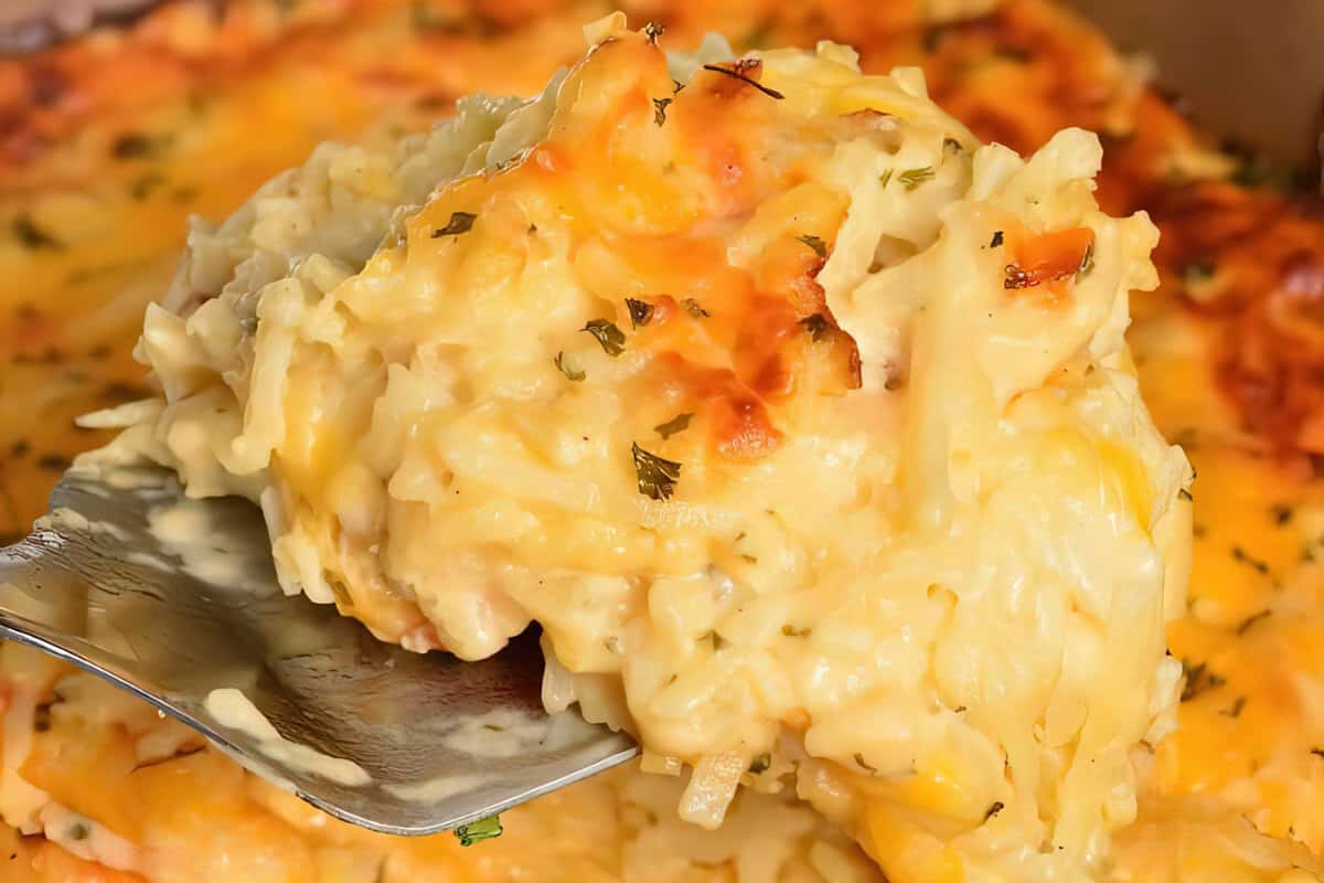 A close-up of a spoon lifting a portion of cheesy hashbrown casserole with melted cheese, herbs, and a golden-brown crust. The casserole appears creamy and well-baked, with grated potatoes and melted cheese visible.