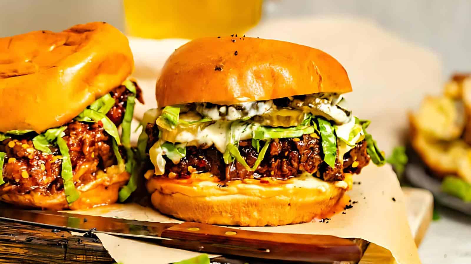 Two juicy burgers with shredded lettuce, pickles, and sauce on toasted buns are shown on parchment paper. A knife is in the foreground and a beverage is in the background.