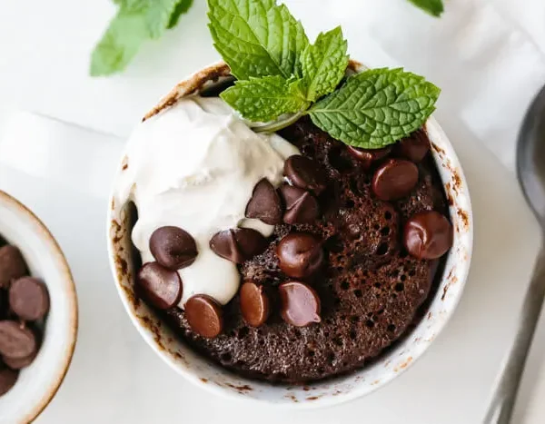 A chocolate mug cake topped with whipped cream, chocolate chips, and mint leaves sits beside a spoon and a small bowl of chocolate chips.