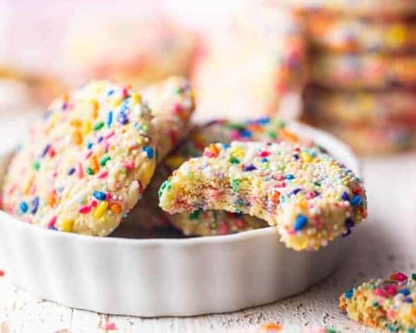A white dish with funfetti-sprinkled cookies, one of which is partially eaten. Additional funfetti cookies appear in the blurred background.