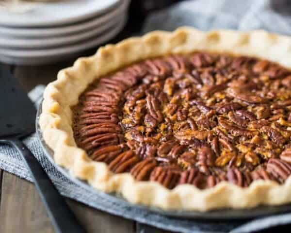 A baked pecan pie on a blue cloth, accompanied by a black pie server and a stack of white plates in the background.