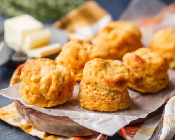Golden-brown biscuits are arranged on wax paper atop a wooden board. In the background, there is a plate with slices of butter and a knife.