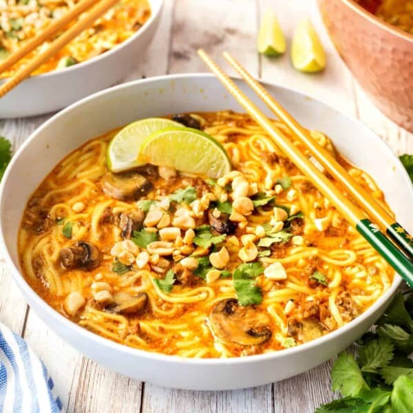 A bowl of ramen with creamy broth, garnished with lime wedges, cilantro, chopped nuts, and mushrooms. Two chopsticks rest on the bowl's edge. Other similarly served bowls are in the background.