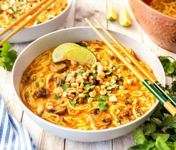 A bowl of noodle soup garnished with lime wedges, chopped peanuts, sliced mushrooms, and fresh herbs, with chopsticks resting on the edge.