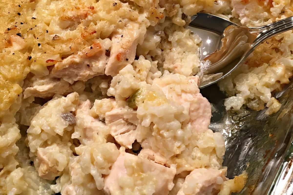 A close-up of a baked chicken and rice casserole in a glass dish. The dish contains chunks of cooked chicken and creamy rice, topped with a slightly browned and crispy layer of cheese. A serving spoon is partially dipped into the casserole.