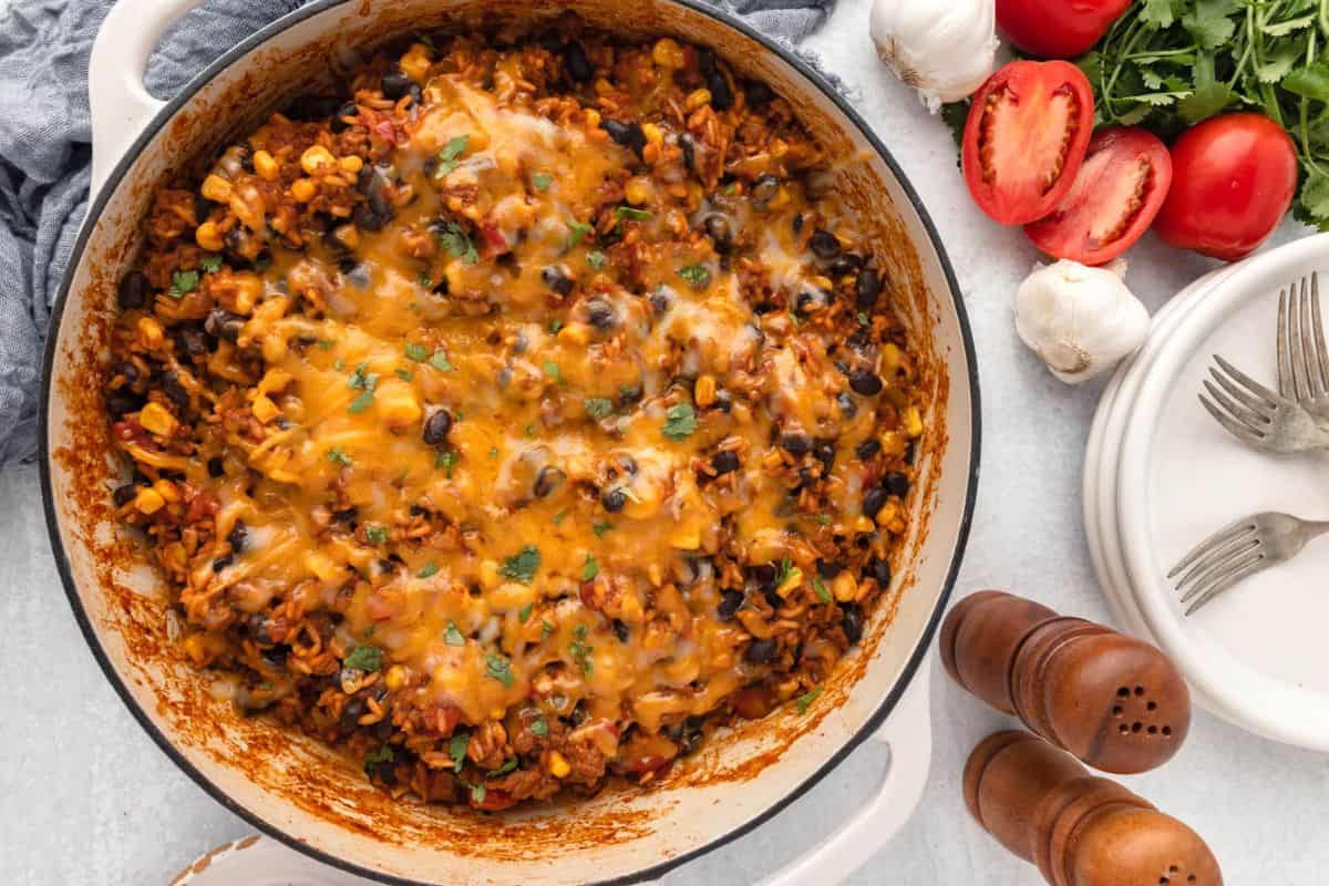 A skillet filled with cheesy rice, black beans, corn, and tomatoes sits on a table next to fresh tomatoes, garlic bulbs, herbs, plates, forks, and salt and pepper shakers.