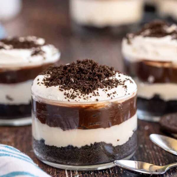 Three glass cups layered with crushed cookies, cream, and chocolate, topped with chocolate crumbs. They sit on a wooden table with a blue striped cloth and three spoons nearby.