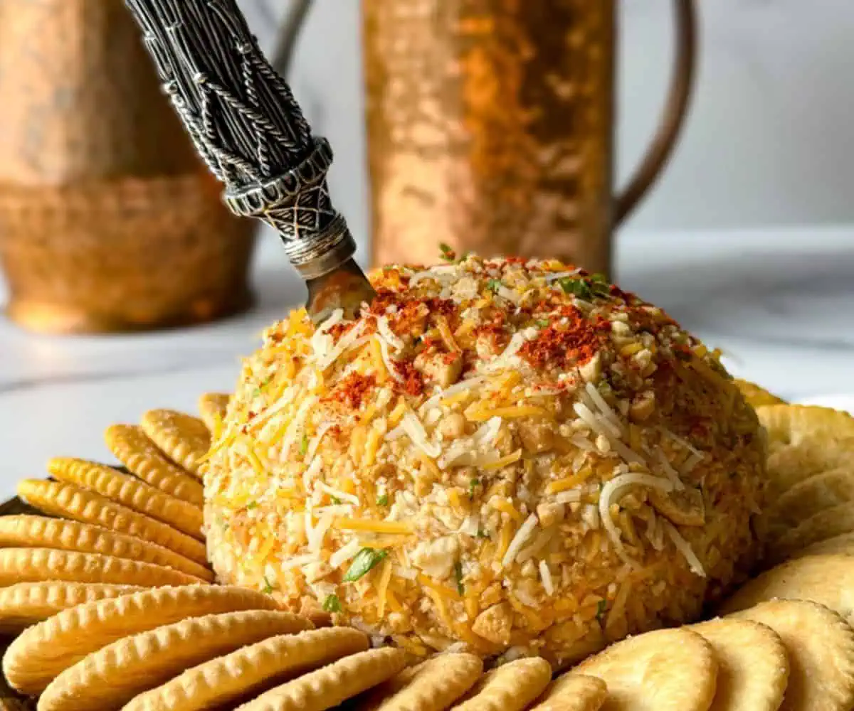 A dome-shaped cheese ball coated with spices and garnished with herbs, surrounded by a ring of circular crackers. A decorative silver knife is inserted into the cheese ball. Copper jugs are blurred in the background.