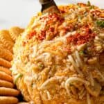 A pineapple cheese ball with shredded cheese and herbs on top is placed on a platter surrounded by round crackers. A spreading knife is inserted into the top of the cheese ball.