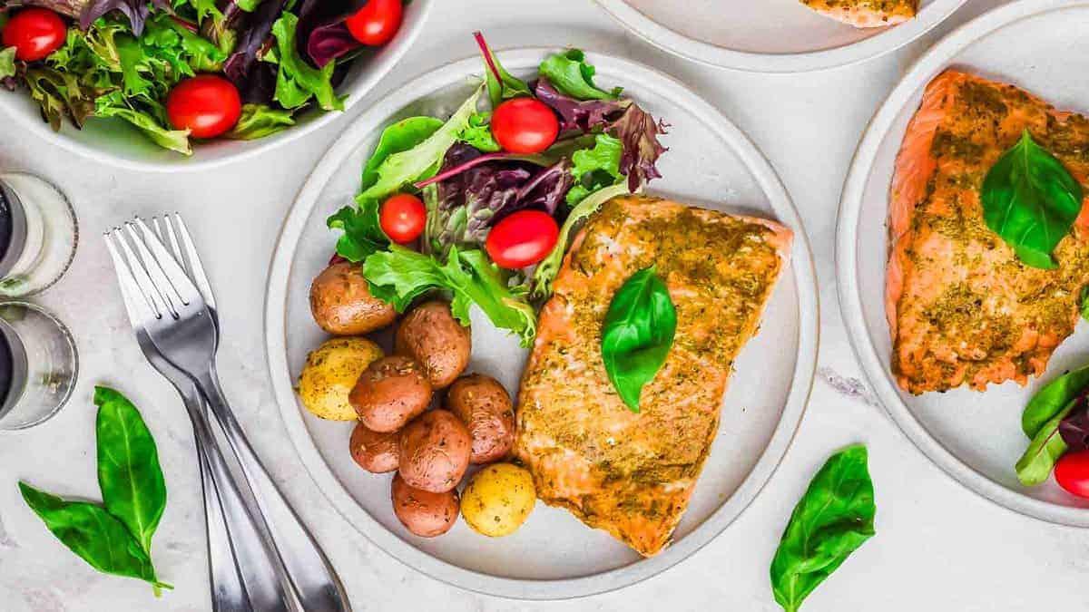 A plate of roasted salmon topped with basil, served with mixed greens, cherry tomatoes, and baby potatoes. Two identical plates and forks are visible nearby.