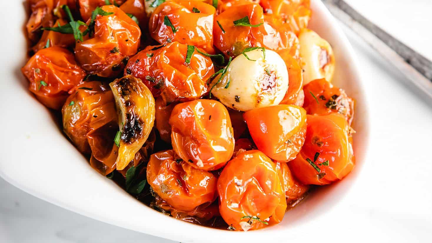A white bowl filled with roasted cherry tomatoes and garlic cloves, garnished with chopped herbs.