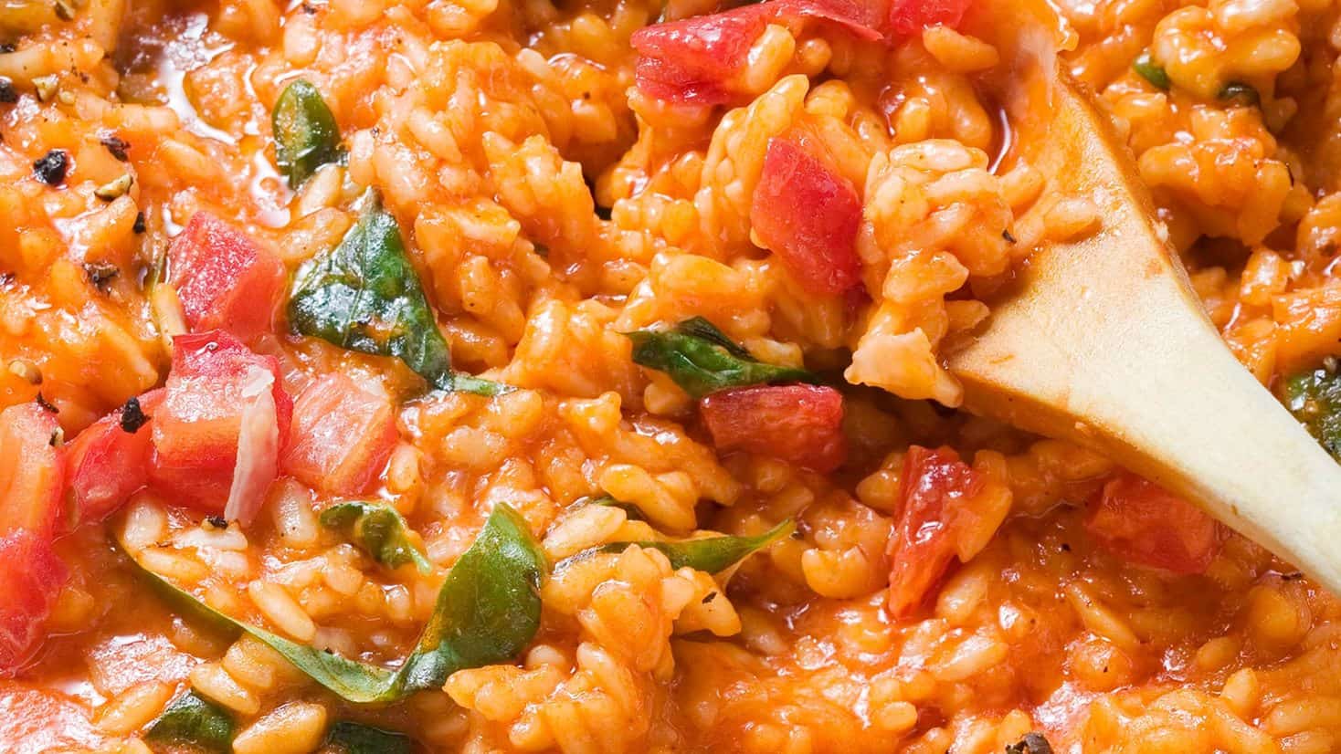 Wooden spoon stirring creamy risotto with visible chunks of tomatoes and green leafy vegetables.
