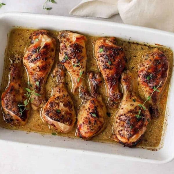 A white baking dish with seven seasoned, baked chicken drumsticks garnished with fresh herb sprigs.
