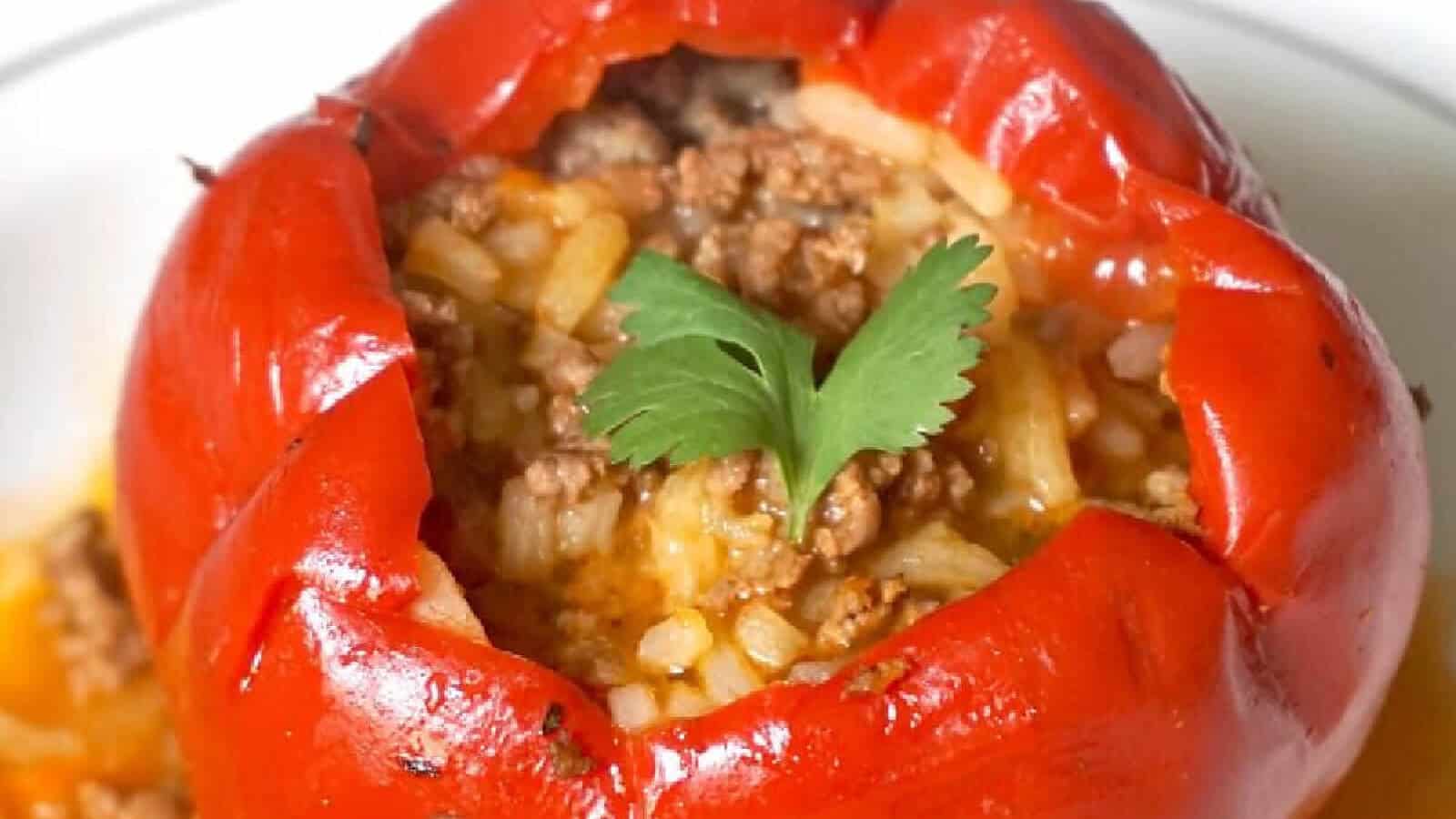 A red bell pepper stuffed with cooked rice and seasoned ground beef, topped with a cilantro leaf, on a pristine white plate.