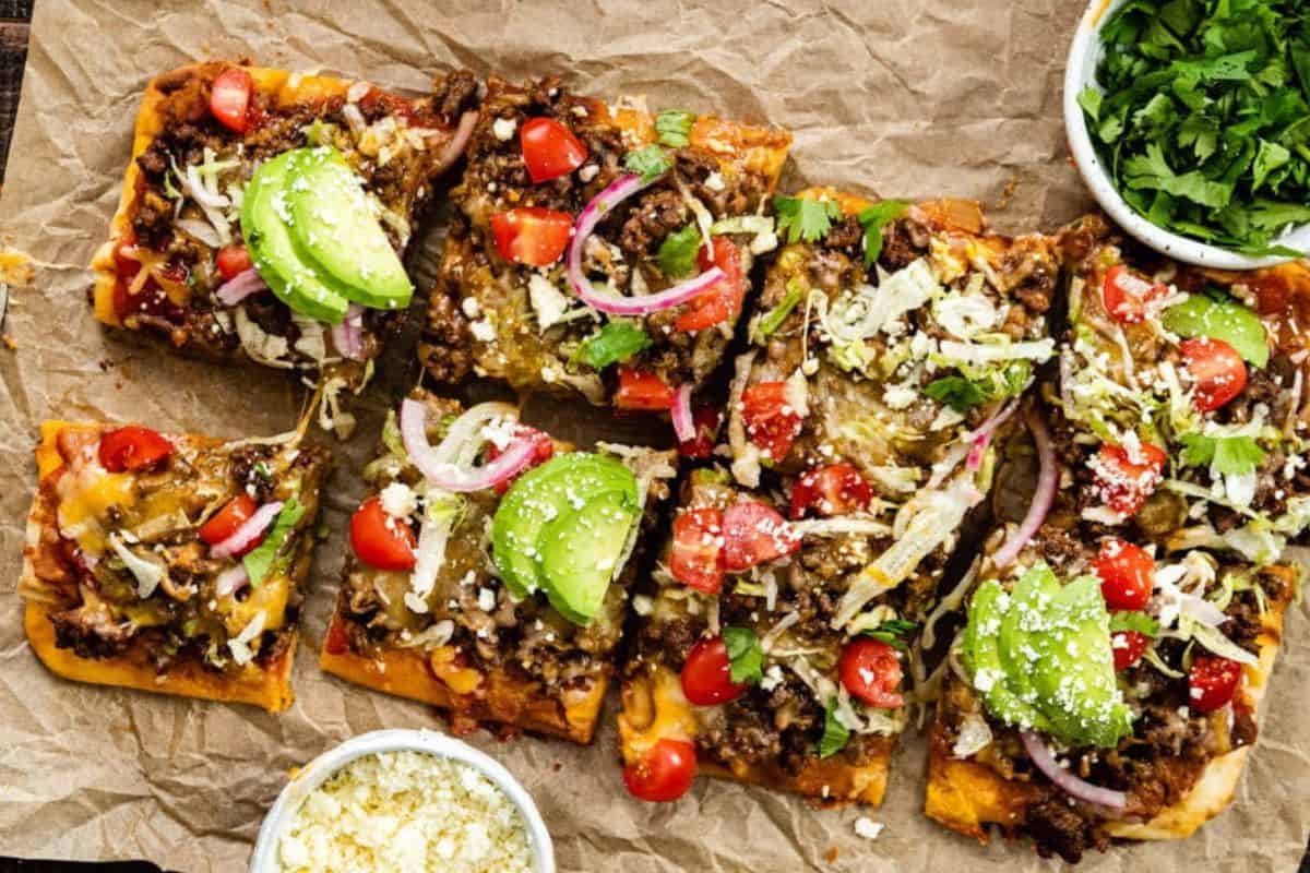 Rectangular pieces of taco pizza topped with ground beef, melted cheese, sliced avocado, halved cherry tomatoes, shredded lettuce, sliced red onion, and chopped cilantro are arranged on parchment paper. Small bowls filled with extra toppings are placed nearby.