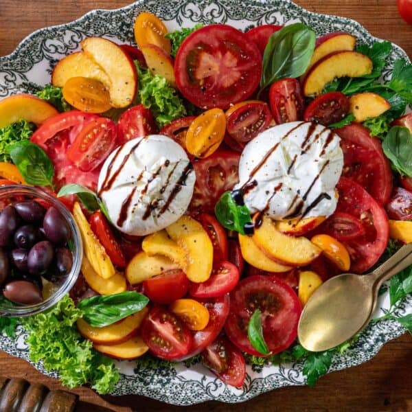 A platter arranged with sliced tomatoes, peaches, and mozzarella is garnished with basil leaves and drizzled with balsamic glaze. Olives are served on the side. Peaches, whole tomatoes, and utensils are placed around the plate.
