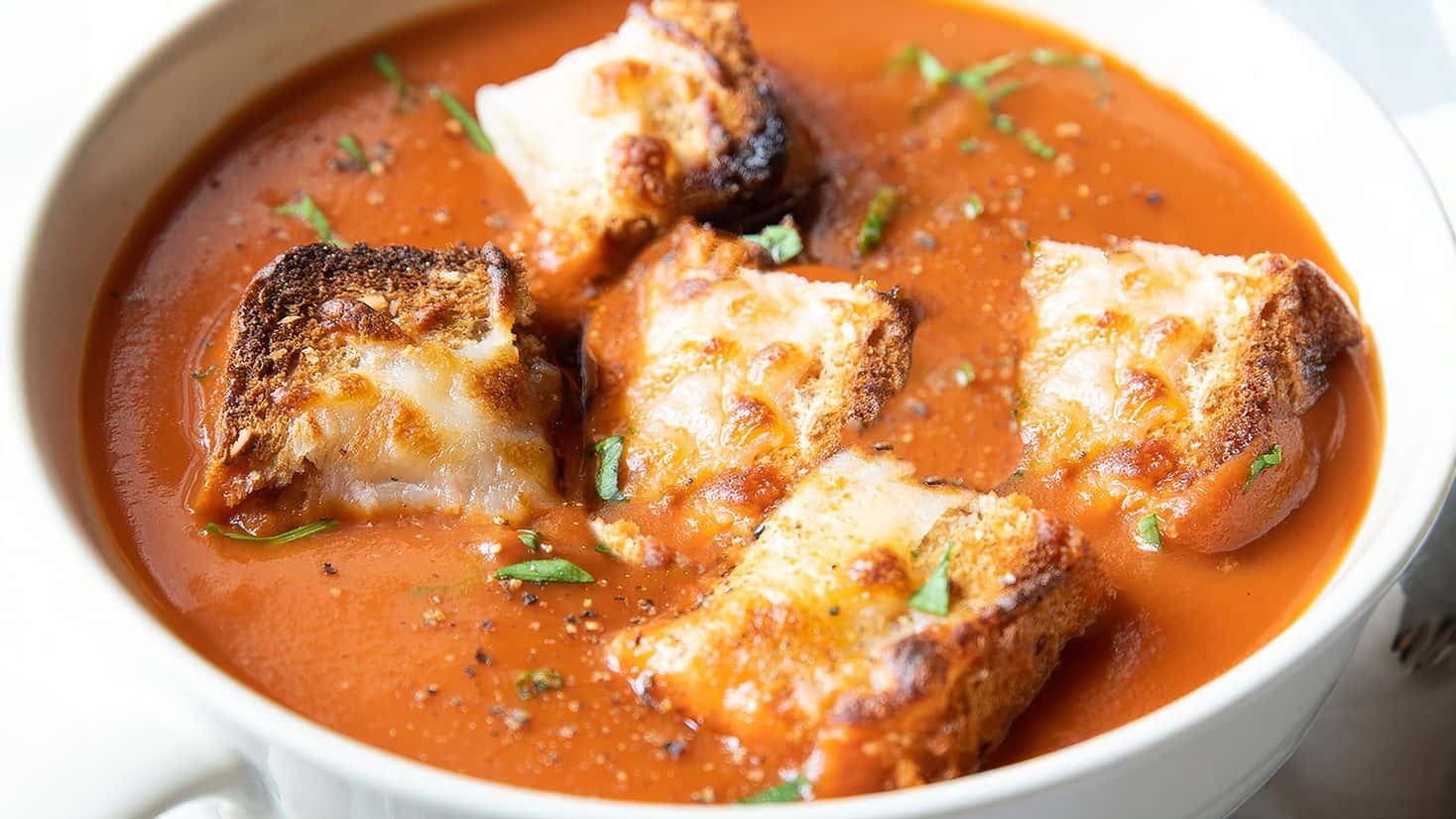 A bowl of tomato soup topped with browned, toasted croutons and garnished with small green herbs.