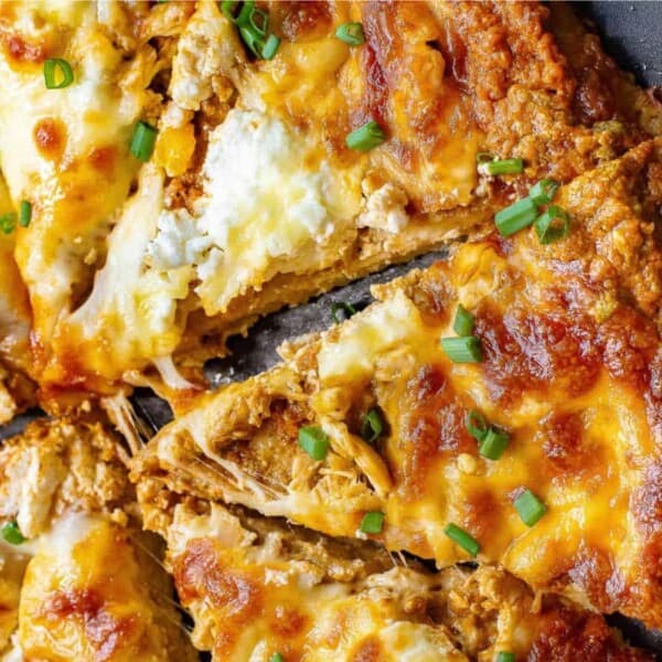 A close-up of a sliced pizza with melted cheese, shredded chicken, and chopped green onions on a dark pan.
