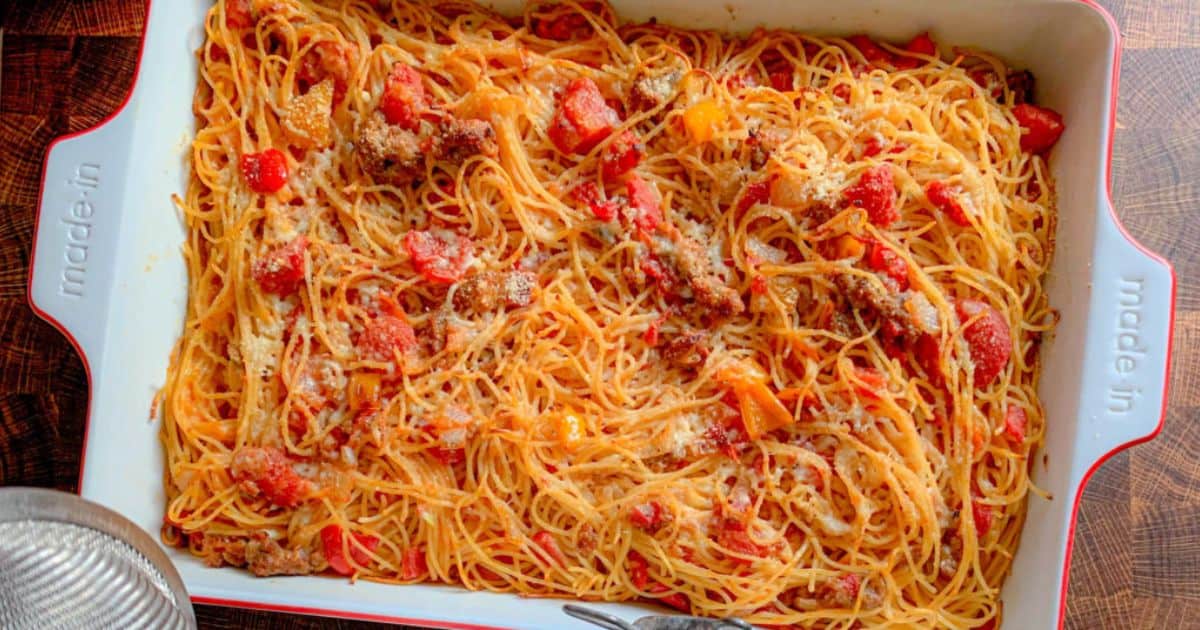 A rectangular baking dish filled with cooked spaghetti, ground meat, diced tomatoes, and bell peppers, sprinkled with grated cheese. A fork and a drinking glass are partially visible nearby.
