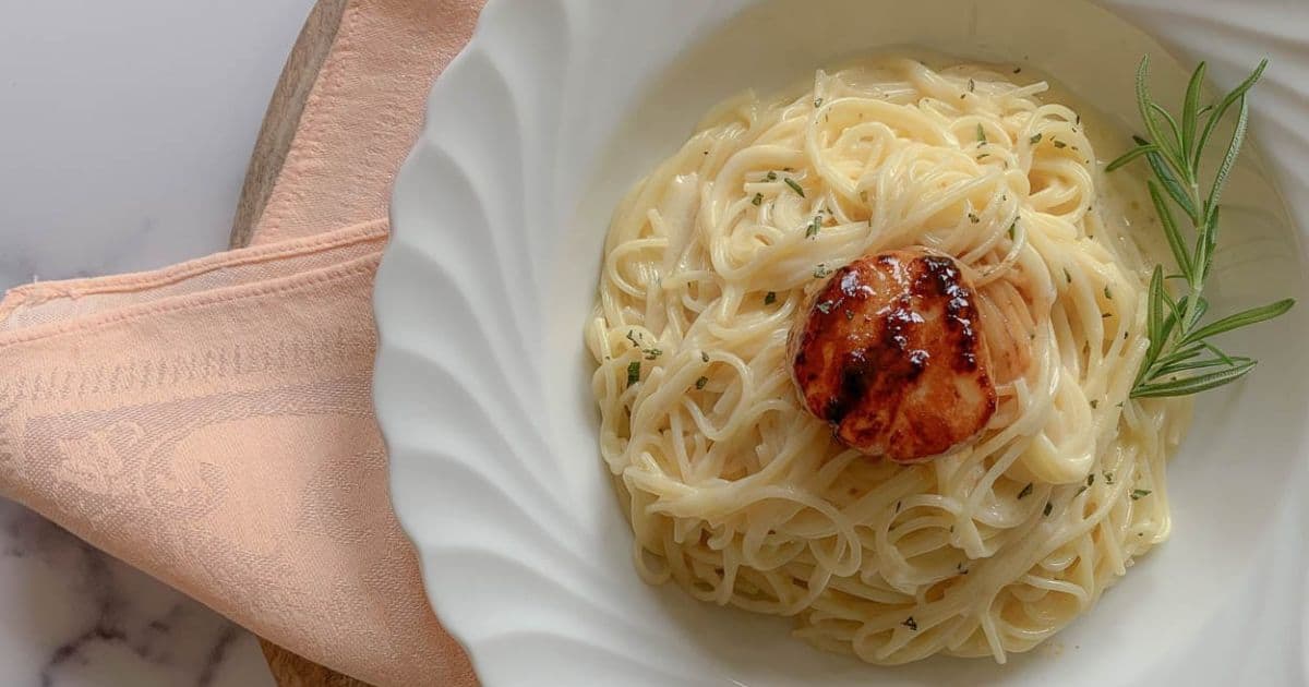 A white plate with creamy pasta topped with a grilled scallop and garnished with a sprig of rosemary, set next to a peach-colored napkin.