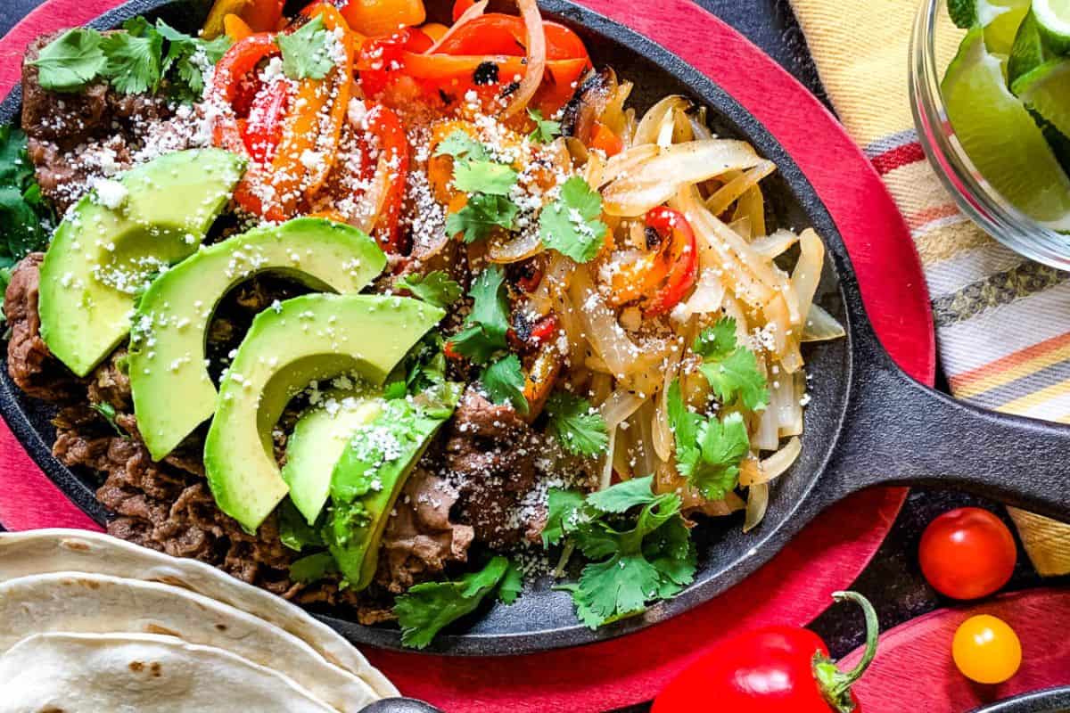 A skillet containing sliced avocado, sautéed onions and peppers, refried beans, fresh cilantro, and crumbled cheese. Tortillas and lime wedges are served on the side.