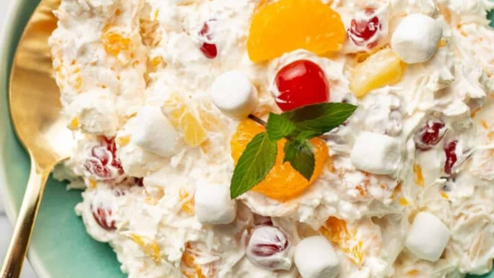 A plate of ambrosia salad with marshmallows, mandarin orange segments, cherries, pineapple, and coconut, garnished with a sprig of mint next to a golden spoon.