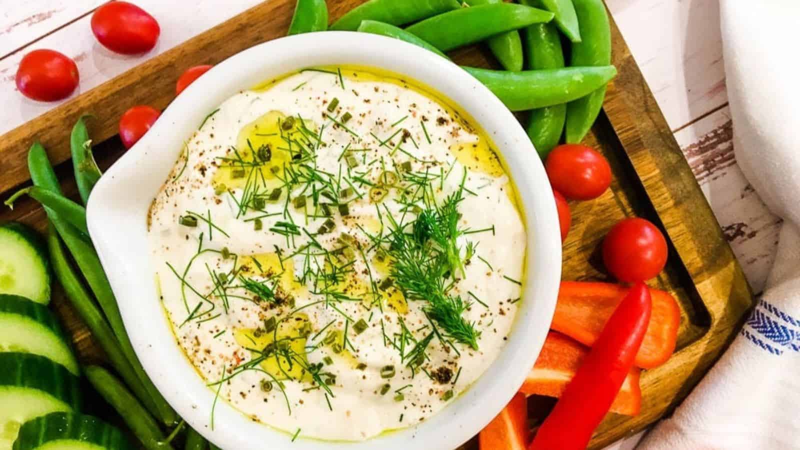 A bowl of creamy dip garnished with herbs and spices, surrounded by cherry tomatoes, green beans, sugar snap peas, cucumber slices, and red bell pepper strips on a wooden tray.