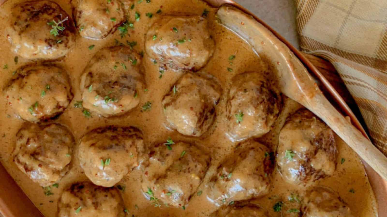 A dish of Swedish meatballs covered in creamy brown gravy, garnished with chopped parsley, sits in a baking dish next to a plaid cloth.