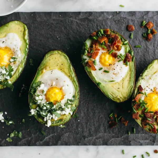Four halved avocados are baked with eggs, garnished with chopped herbs, crumbled cheese, and diced bacon, all arranged on a dark slate surface. A portion of one avocado half is partially visible, along with scattered garnish around them.